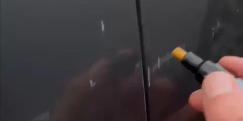 Eine Hand trägt einen Klarlack-Reparaturstift direkt auf einen Autokratzer auf.